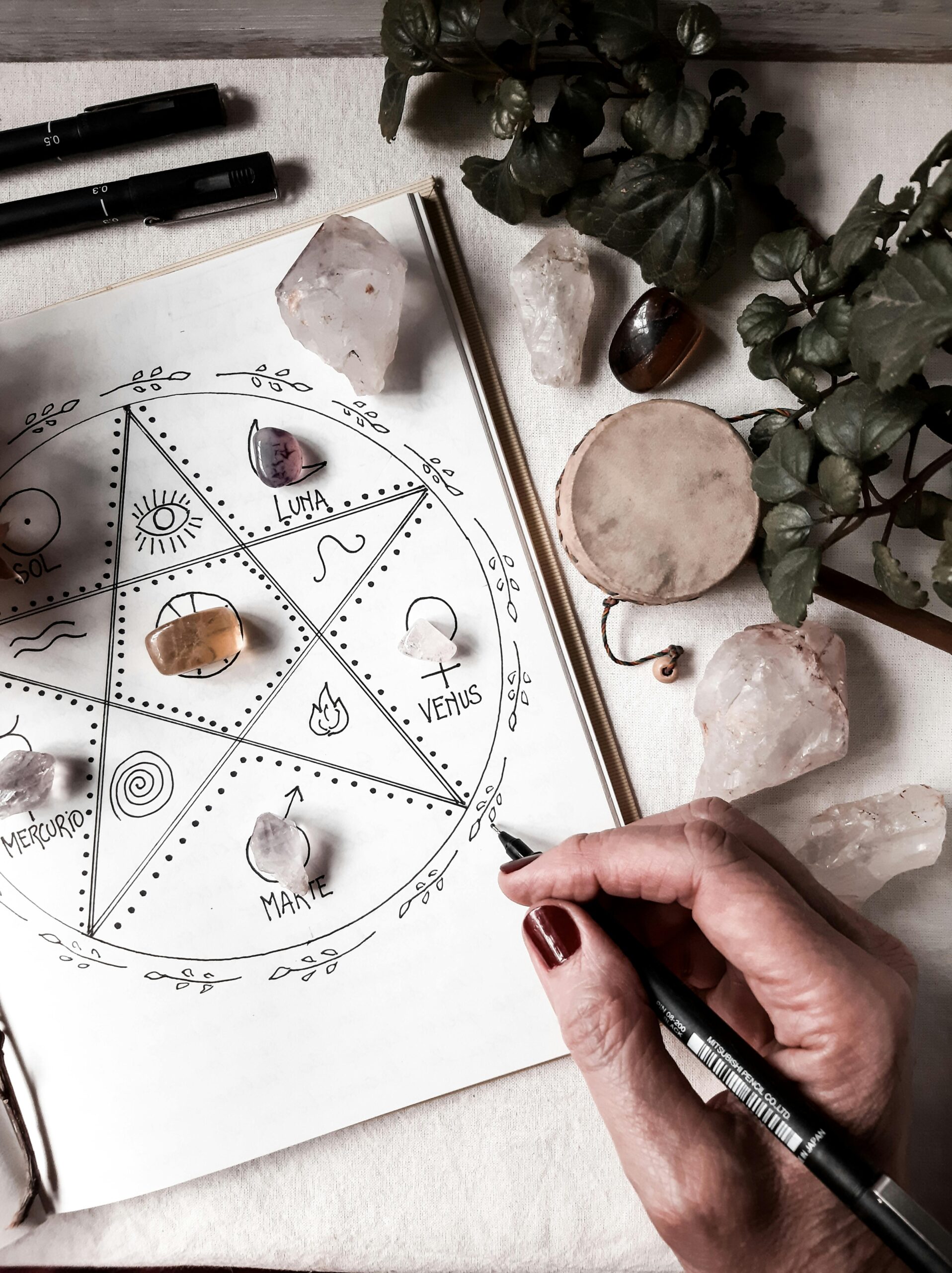 A hand drawing an astrology chart surrounded by crystals and plants, capturing a mystical atmosphere.